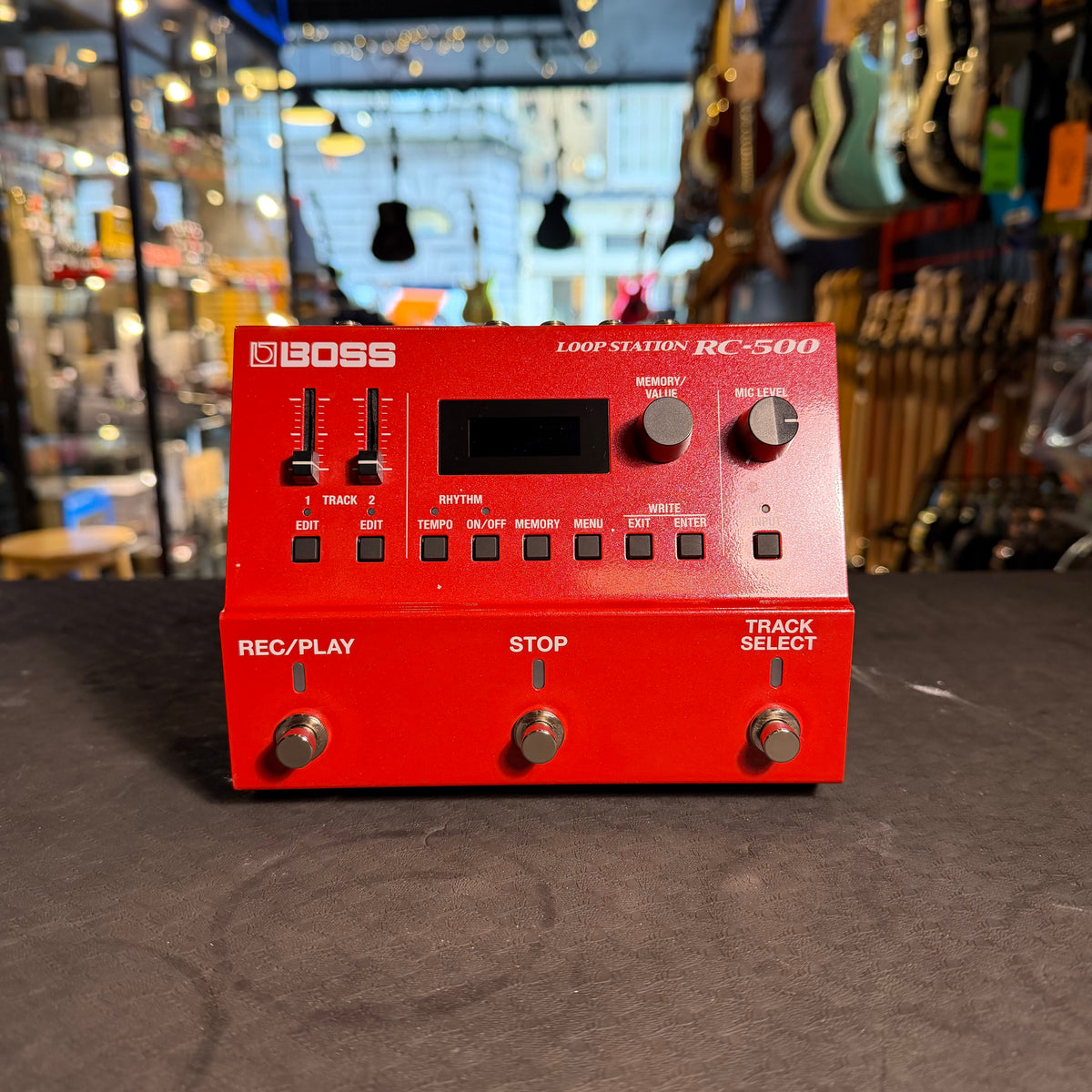Red BOSS Loop Station RC-500 pedal on a dark surface with a guitar store background