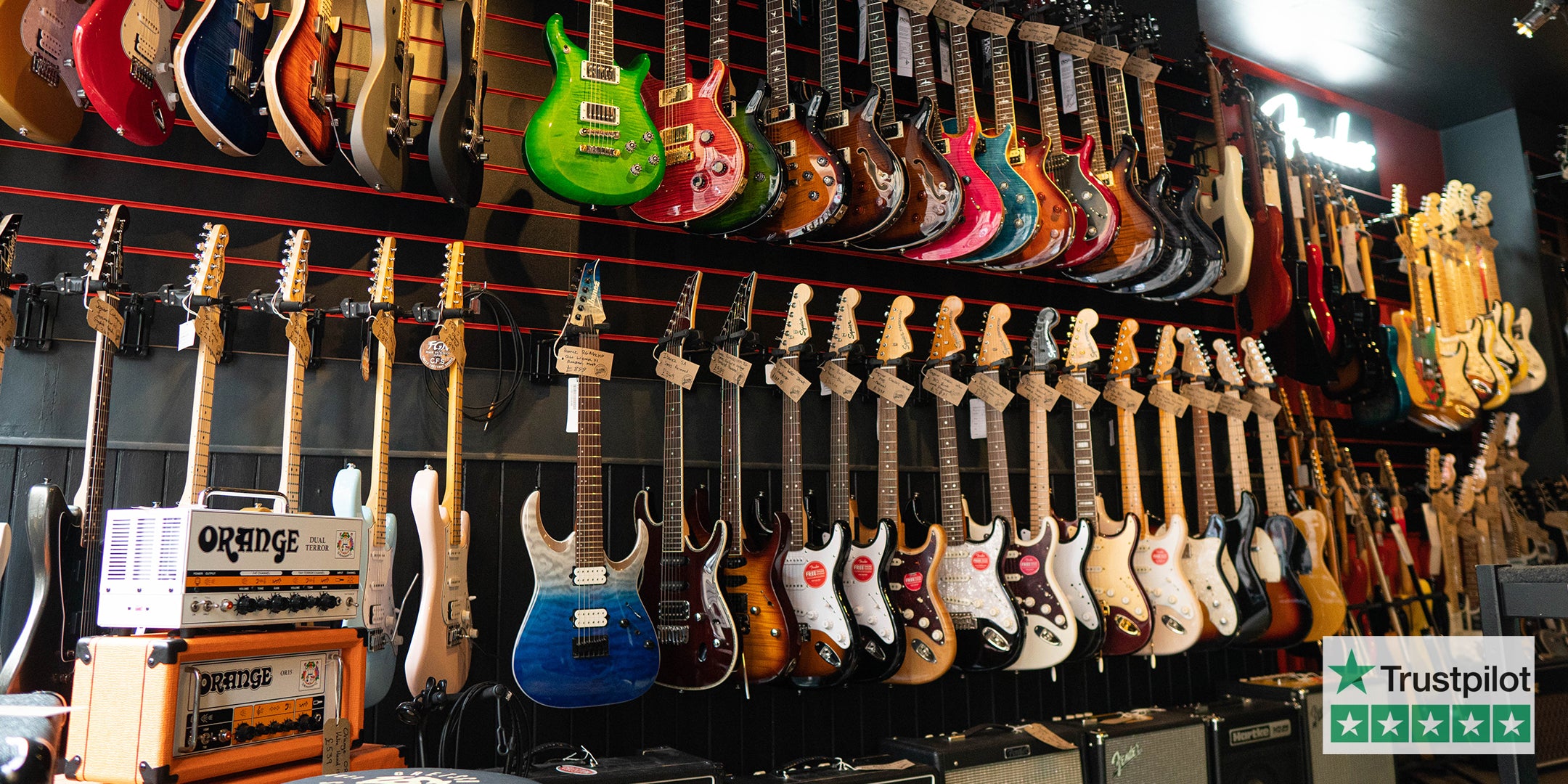 2 racks of electric guitars of all colours hanging on the wall surrounded by Amps at Guitar Bitz Guitar Shop